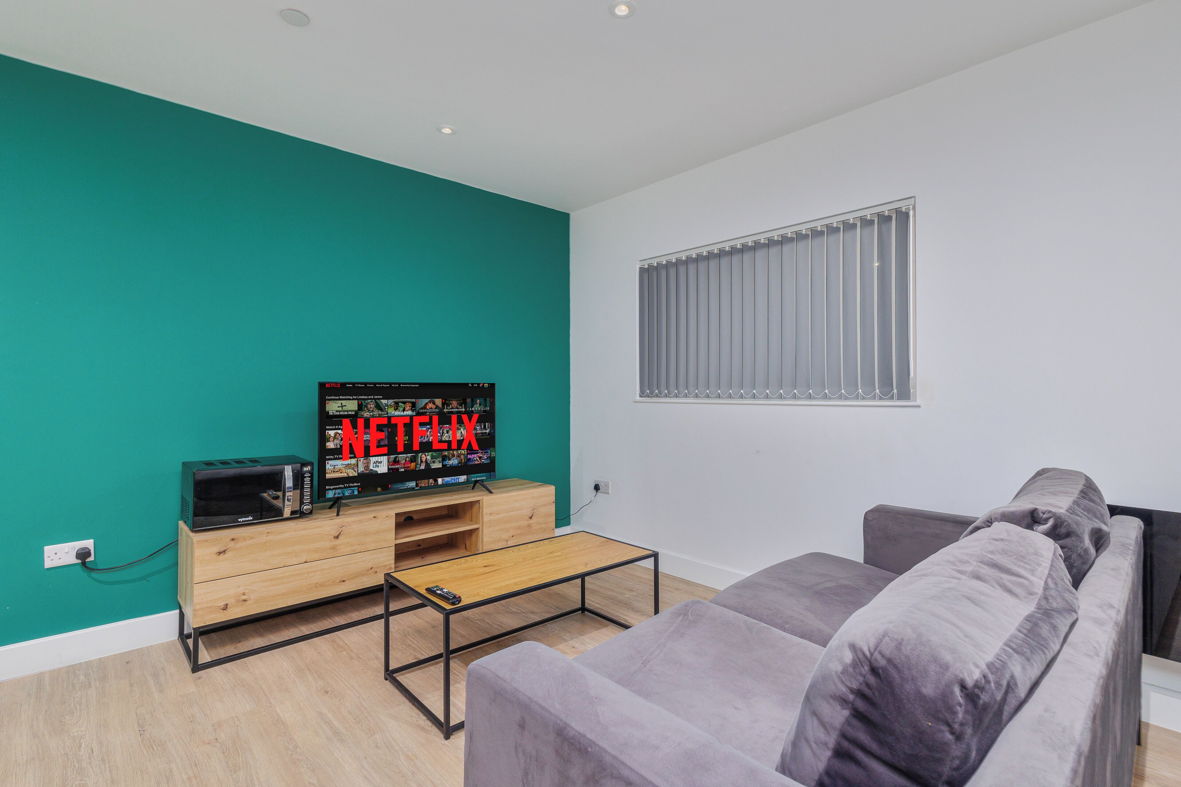 A modern living room with teal walls, a grey sofa, a wooden TV stand, and a large window with blinds.