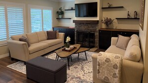 A living room with a sofa, ottoman, coffee table, fireplace, TV, and decorative shelves. - Clean, Bright & Comfortable Pool Home w/separate Poolhouse Studio (Los Angeles)
