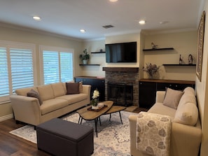 A living room with a sofa, ottoman, coffee table, fireplace, TV, and decorative shelves. - Clean, Bright & Comfortable Pool Home w/separate Poolhouse Studio (Los Angeles)