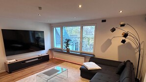 Uma sala de estar moderna com uma grande televisão de tela plana, um sofá escuro, uma mesa de centro de vidro e uma planta perto das janelas.