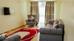 A living room with a sofa, armchair, and a flat-screen TV mounted on the wall. - Ashville Homes (Meru)