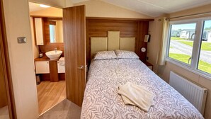 Une chambre à coucher avec une tête de lit en bois, un grand lit recouvert d’une literie à motifs, une table de chevet avec une horloge et une fenêtre donnant sur un quartier résidentiel.