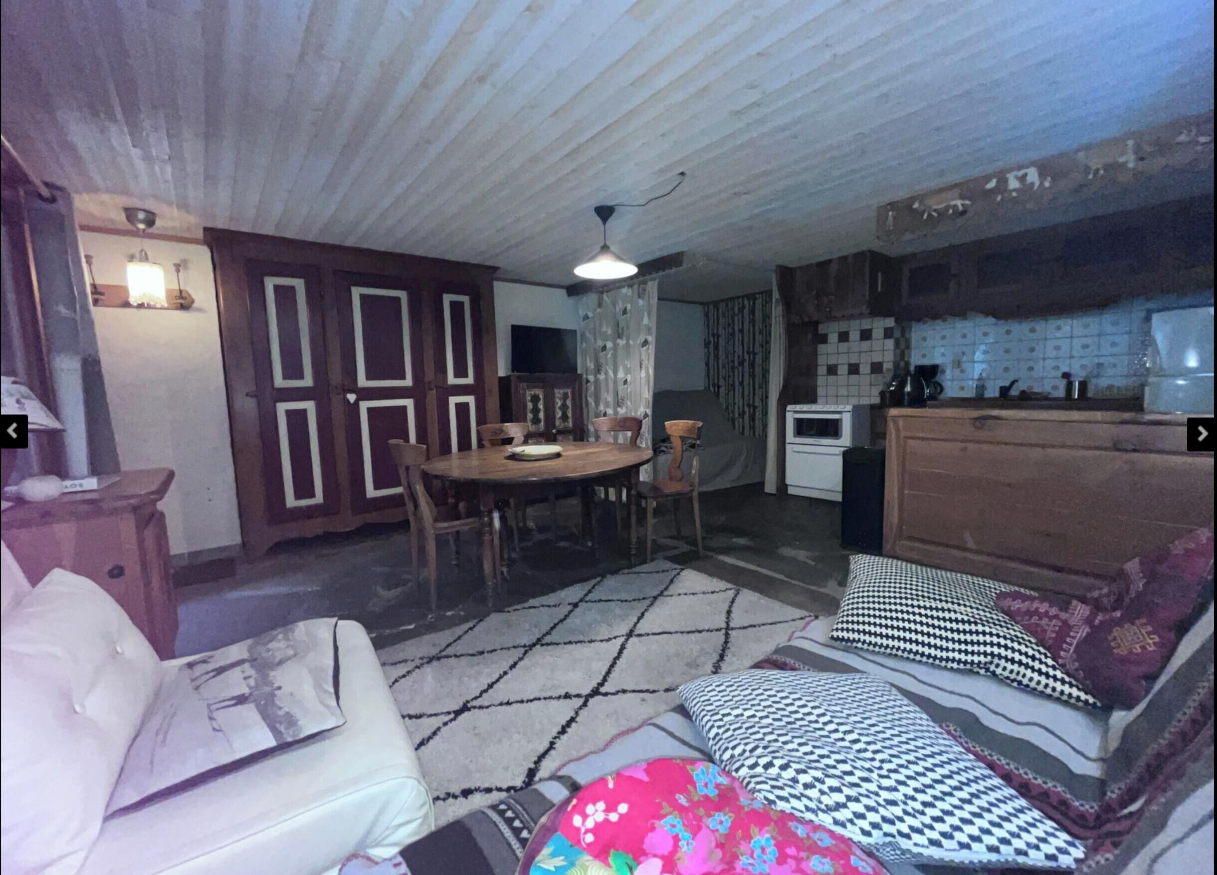 A room with a dining table, chairs, a sofa, a patterned rug, and a kitchenette.