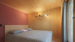 A bedroom with a wooden ceiling, a bed with patterned bedding, a wall-mounted light, and a window with curtains.