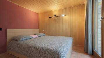 Een slaapkamer met een houten plafond, een bed met beddengoed met een patroon, een wandlamp en een raam met gordijnen.