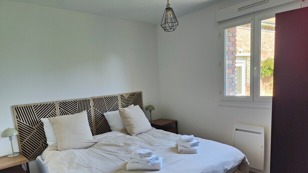 A neatly made bed with pillows and folded towels, a bedside table with a lamp, and a window with a view of a brick building.