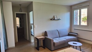 A living room with a grey sofa, a small round table, a wooden coffee table, a hanging light fixture, and a window with a view of a brick building.