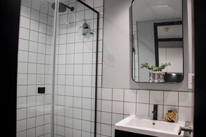 Ein Badezimmer mit Duschkabine aus Glas, einem weißen Waschbecken und einem Spiegel mit Pflanze.