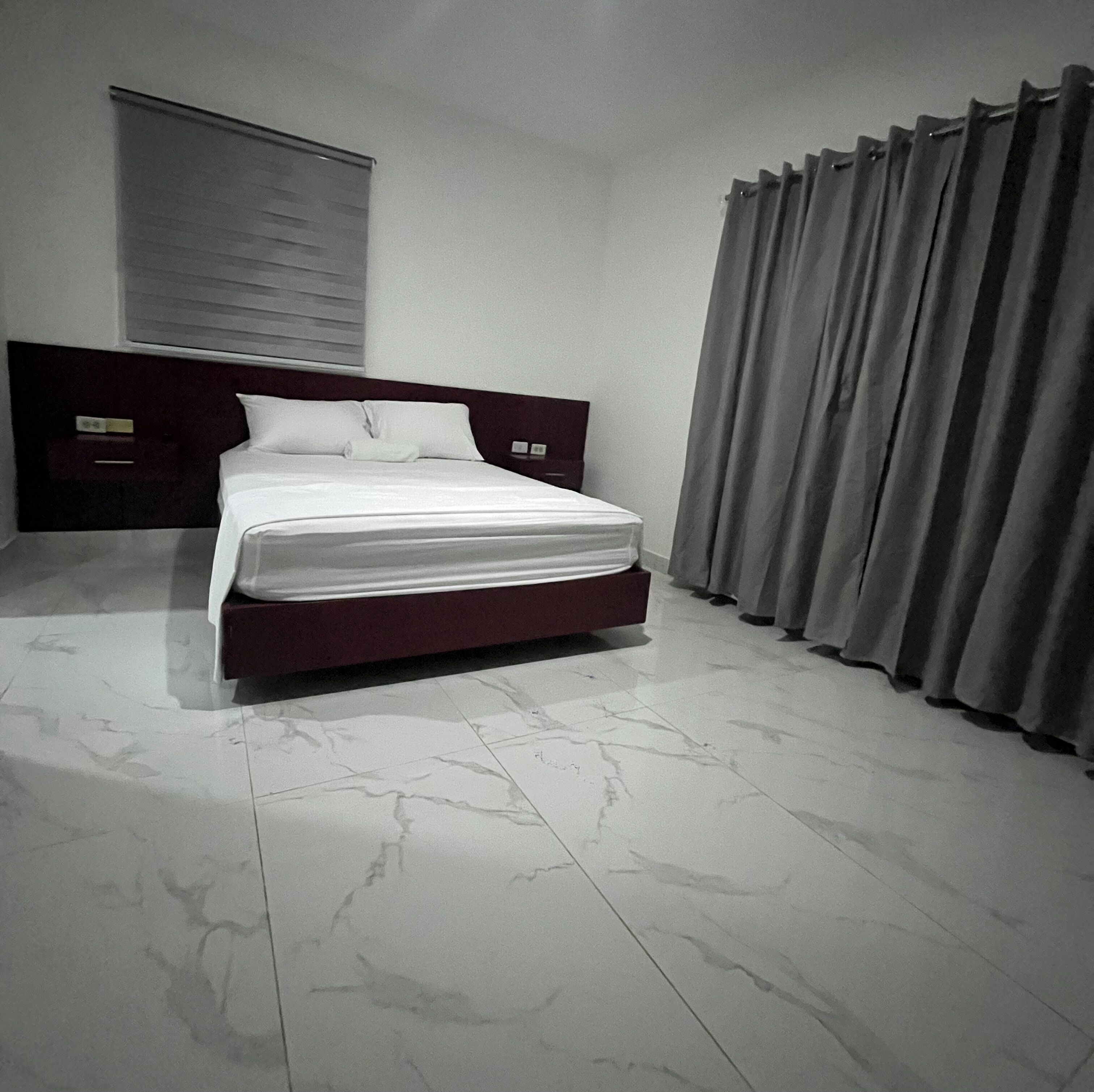 A bedroom with a bed, a headboard, a window with blinds, and a curtain.