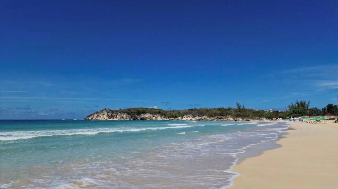 A beach with clear blue water, sandy shore, and a rocky outcrop in the distance.