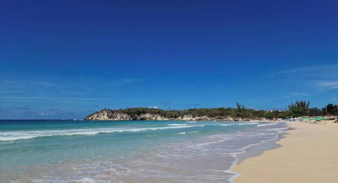 Una playa con agua azul cristalina, orilla de arena y un promontorio rocoso en la distancia.