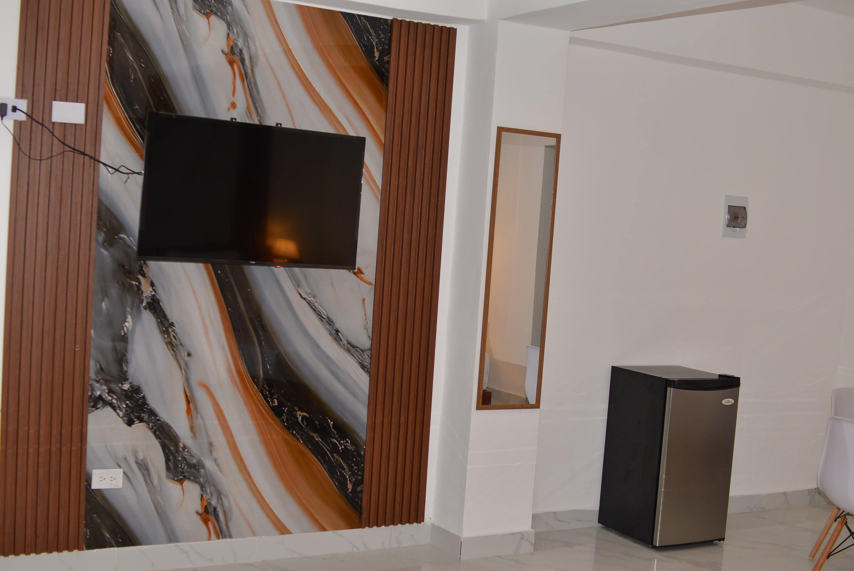A room with a flat-screen TV mounted on a wall with a marble-like design, a small trash bin, and a mirror on the wall.