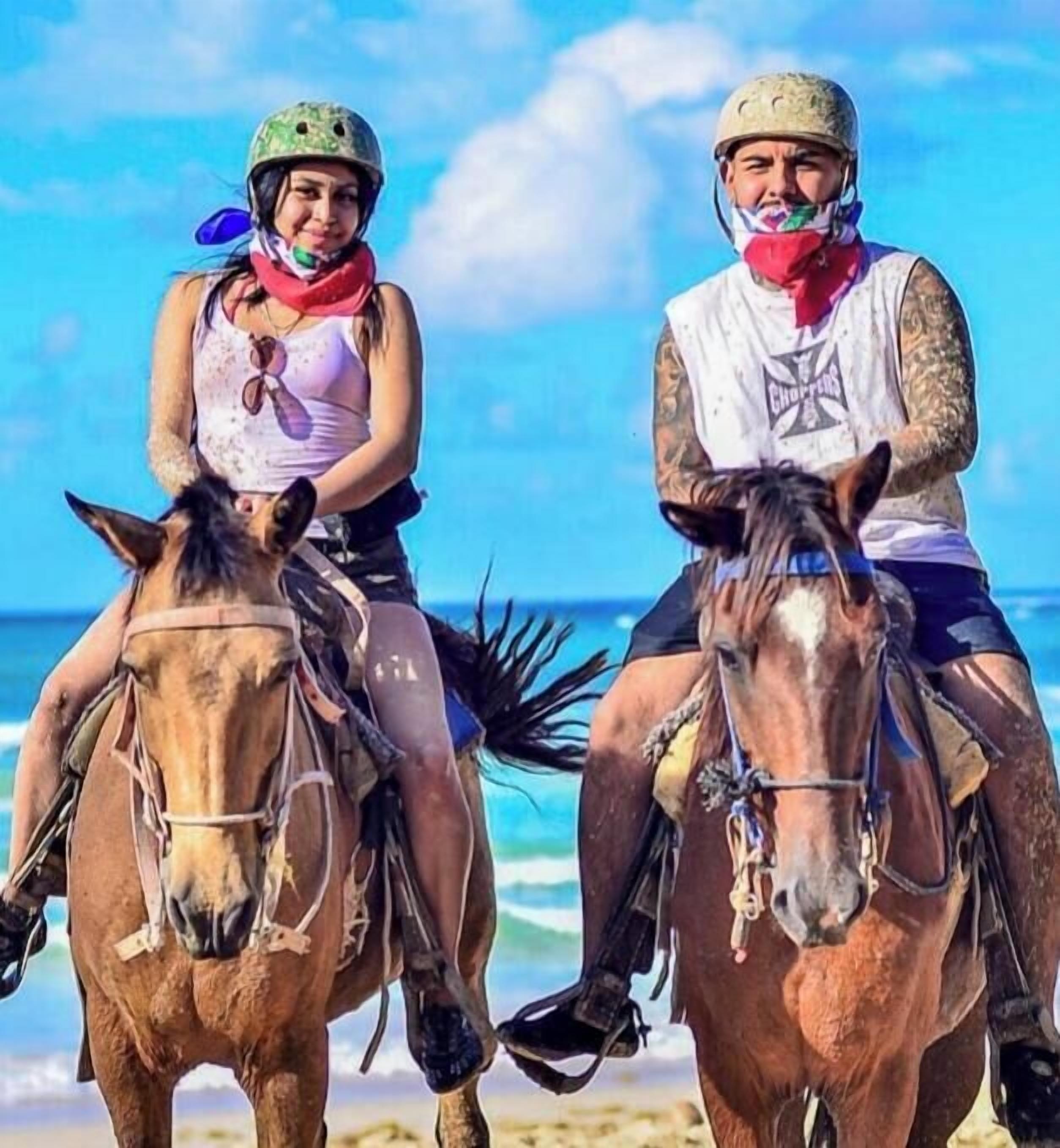 Two people riding horses on a beach with the ocean and a rocky cliff in the background.