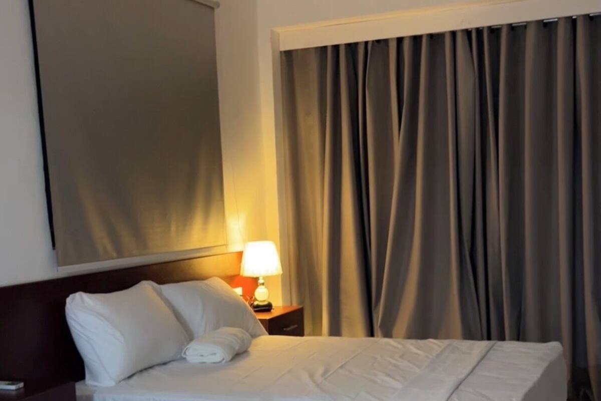 A hotel room with two beds, a nightstand with a lamp, a large window with curtains, and a wall-mounted TV.