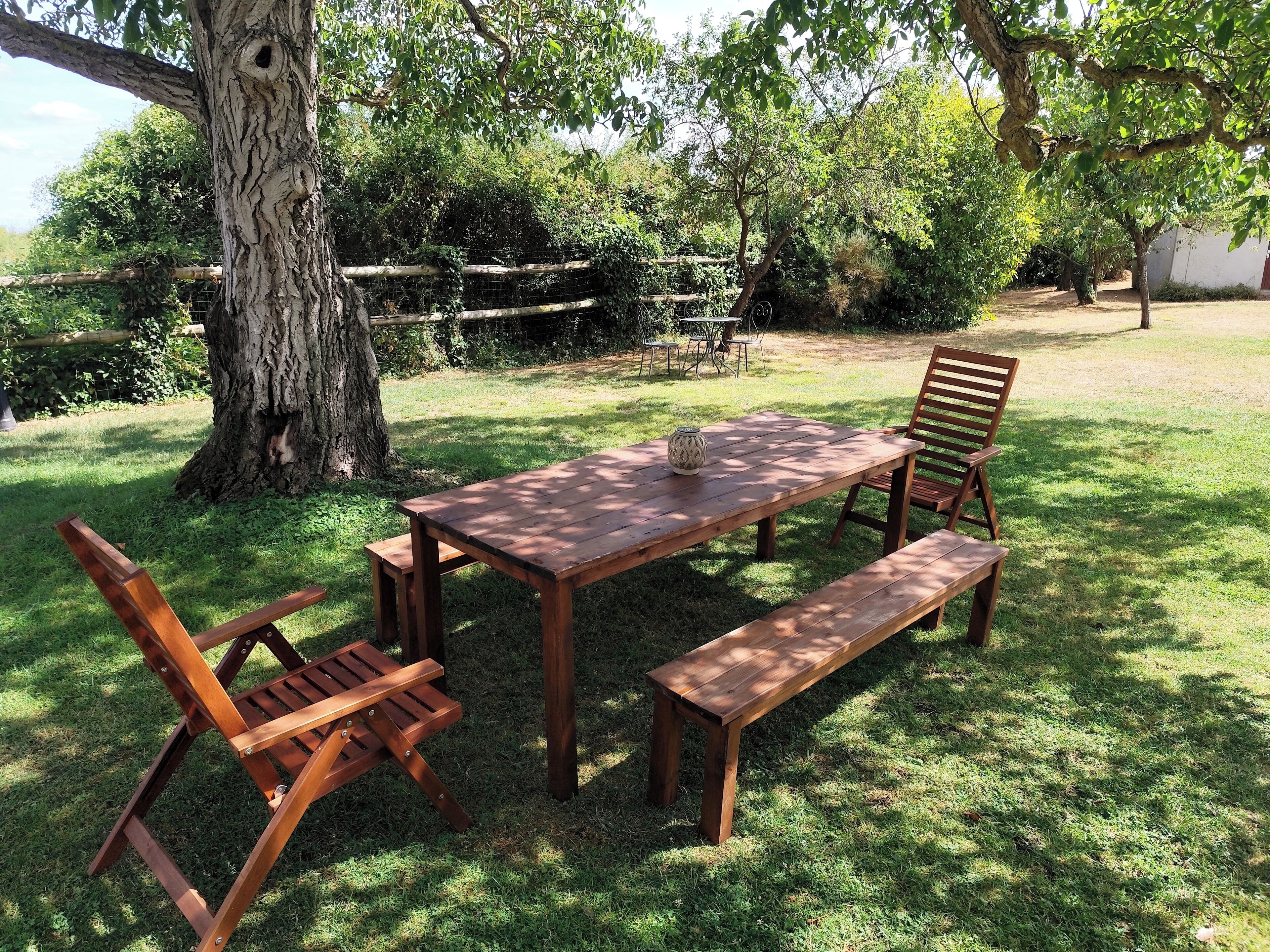 Un conjunto de mesa y bancos de madera al aire libre, sobre un césped con árboles al fondo.
