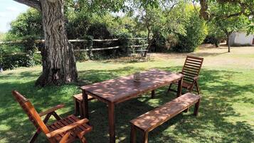 Un conjunto de mesa y bancos de madera al aire libre, sobre un césped con árboles al fondo.