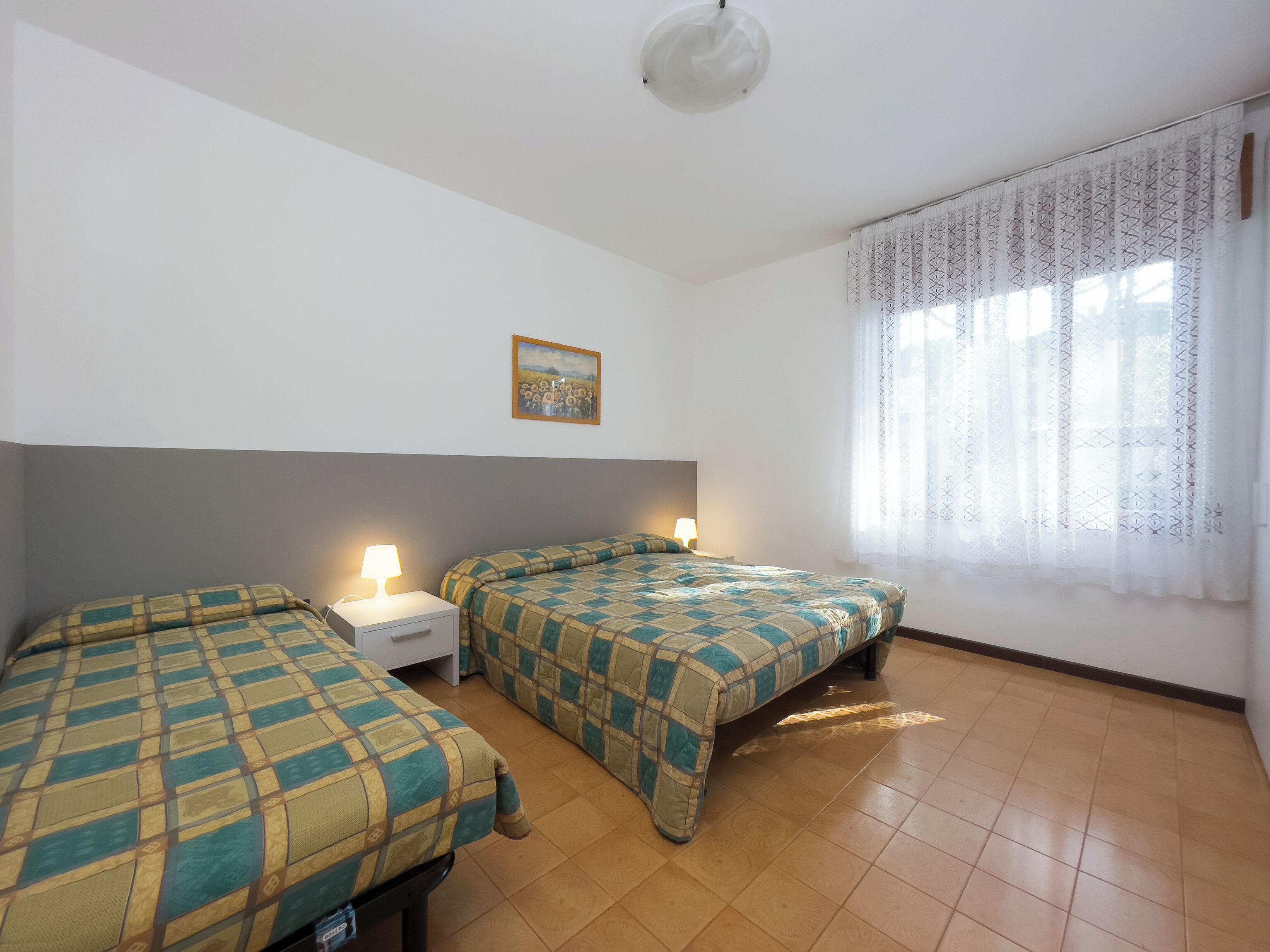 Una habitación con dos camas, una mesita de noche, una lámpara, un cuadro y una ventana con cortinas.
