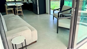 Un espace de vie moderne comprenant un canapé, une petite table ronde, une cuisine avec un réfrigérateur et un balcon offrant une vue sur la verdure.