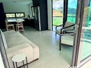 A modern living area with a sofa, a small round table, a kitchen with a refrigerator, and a balcony with a view of greenery. - @NEW Modern El Yunque Rainforest Best View House with Pool@ (Río Grande)
