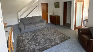 Un salón espacioso con un sofá gris, un sillón de cuero marrón, un tapete de pelo alto color gris y una escalera que sube a otro nivel.