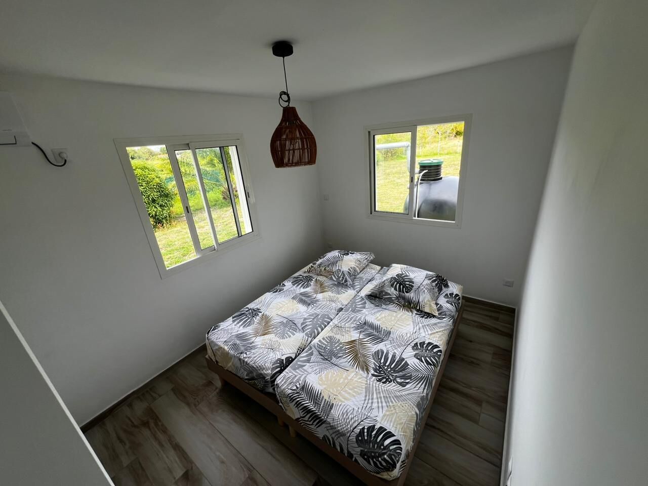 Een slaapkamer met een bed, twee ramen, een hanglamp en uitzicht op groen.
