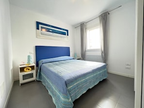 A bedroom with a bed, a nightstand, a wall-mounted picture, and a window with curtains. - Lignano Sabbiadoro Central Flat (Lignano Sabbiadoro)