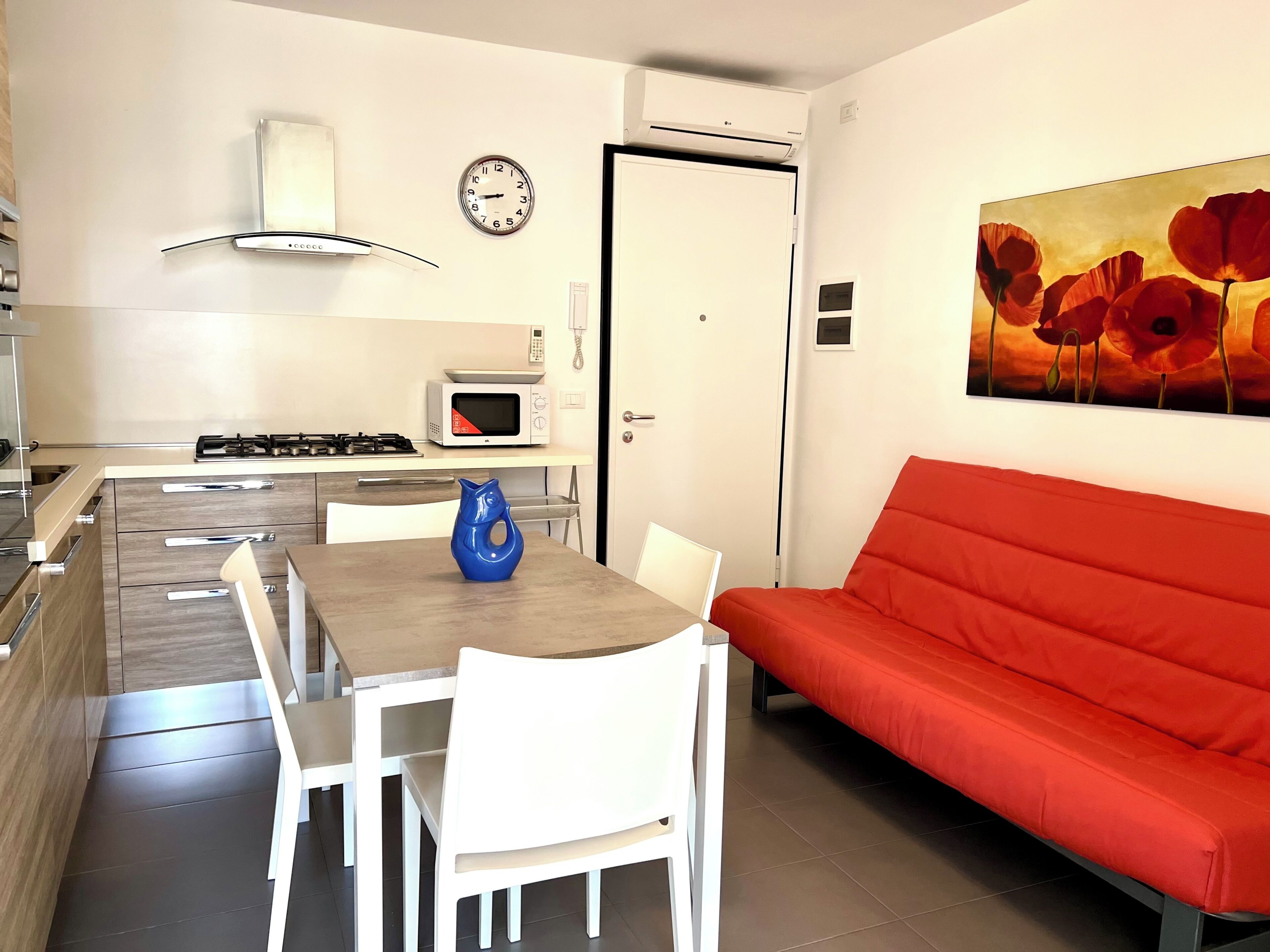 Une cuisine moderne équipée d’un four à micro-ondes, d’une cuisinière et d’une horloge murale. On y trouve une table à manger avec des chaises, ainsi qu’un canapé rouge dans l’espace salon.