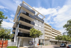A multi-story building with balconies, a green lawn, and parked cars. - Lignano Sabbiadoro Central Flat (Lignano Sabbiadoro)