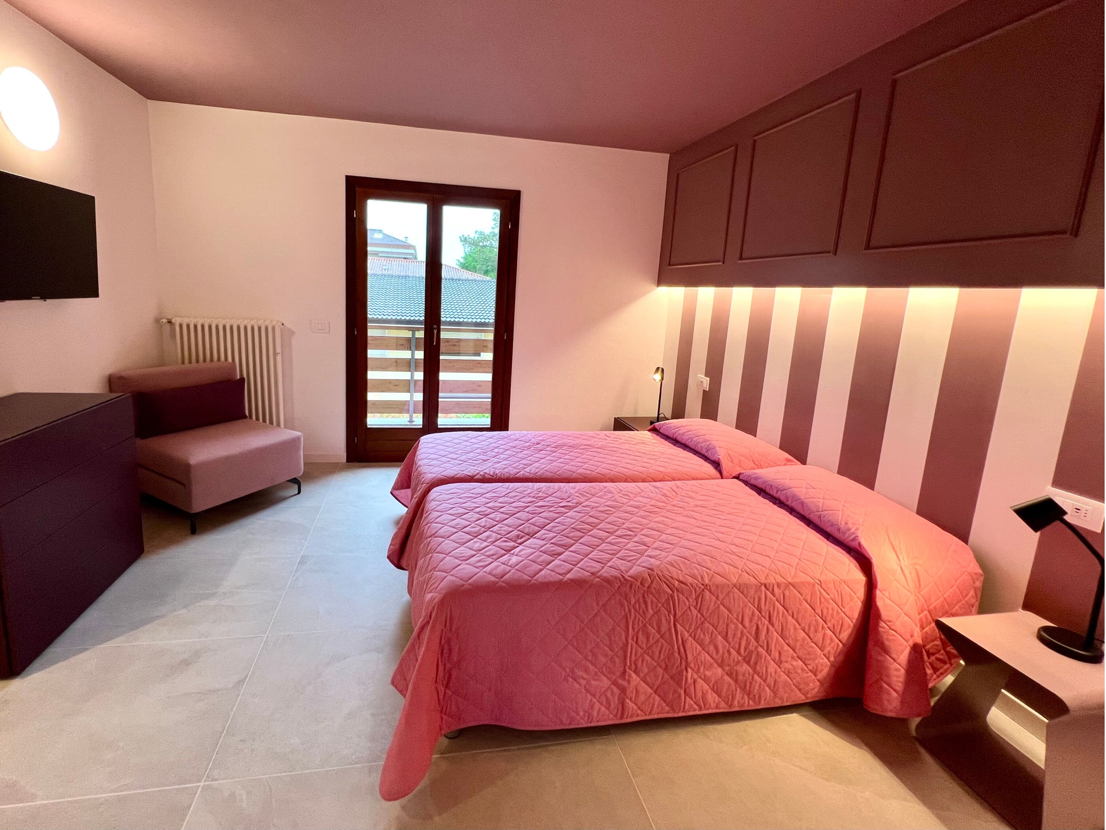 Une chambre avec deux lits, un bureau, une chaise, un radiateur et une porte-fenêtre coulissante donnant sur un espace extérieur.