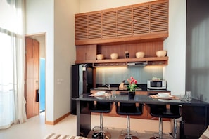 A modern kitchen with a black countertop, three bar stools, a microwave, and a wooden upper shelf with baskets. - Wings 35 Laguna (Phuket)