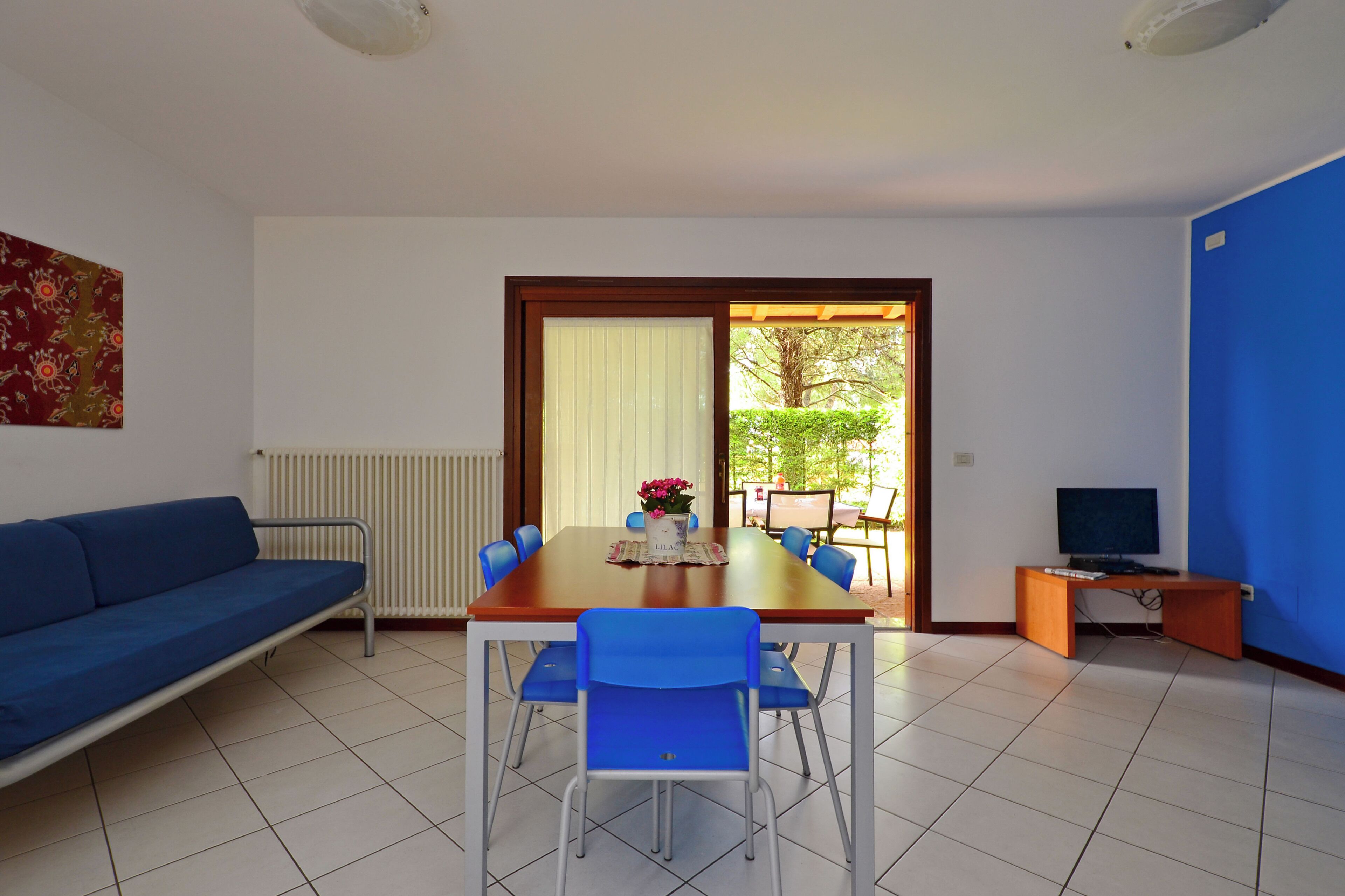 Ein Zimmer mit einem blauen Sofa, einem Esstisch mit Stühlen, einem Fernseher und Blick ins Freie.