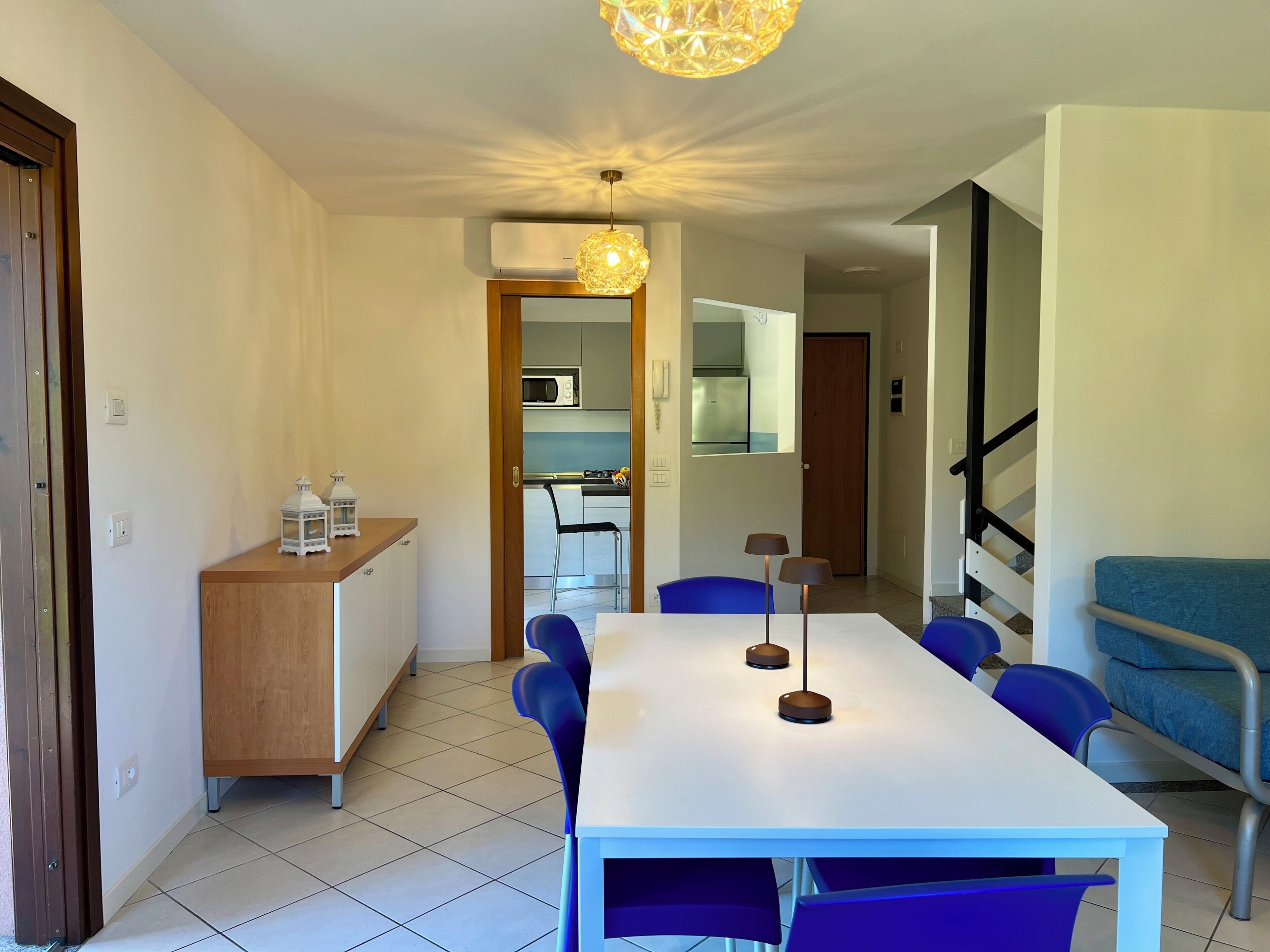 Zona pranzo con tavolo bianco, sedie blu, un buffet in legno e un lampadario.