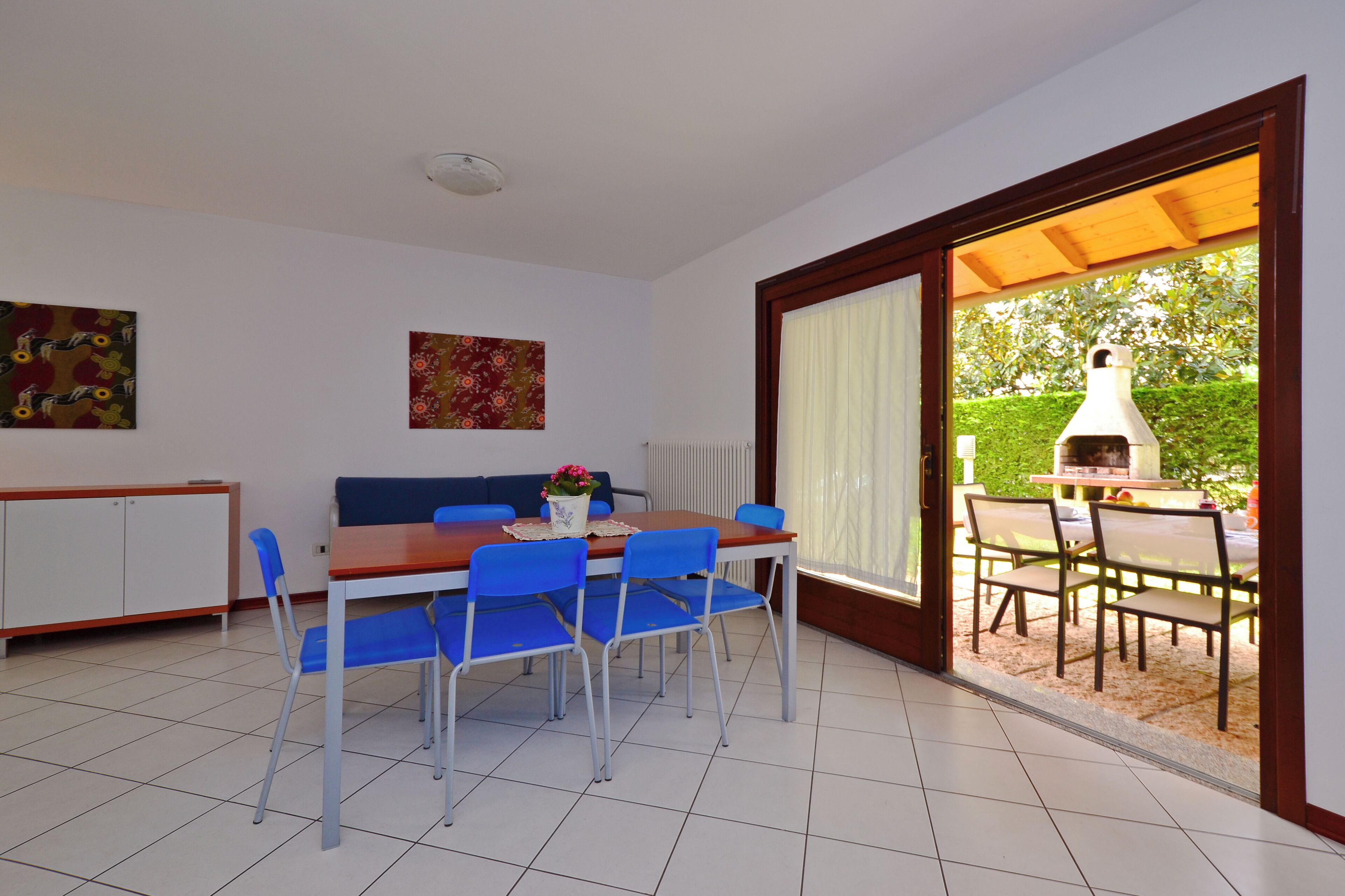 A dining area with a table and chairs, a sofa, and a view of an outdoor barbecue area through a sliding glass door.