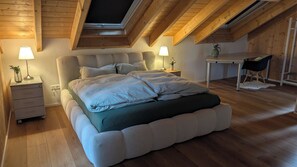 Ein Schlafzimmer mit hohen Decken, einem Bett mit grüner Bettdecke, zwei Nachttischlampen, einem kleinen Tisch und einem Stuhl.