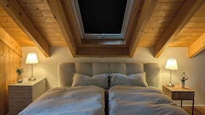 A bedroom with a sloped ceiling, a skylight, two bedside tables with lamps, a bed with striped bedding, and a small nightstand with a vase.