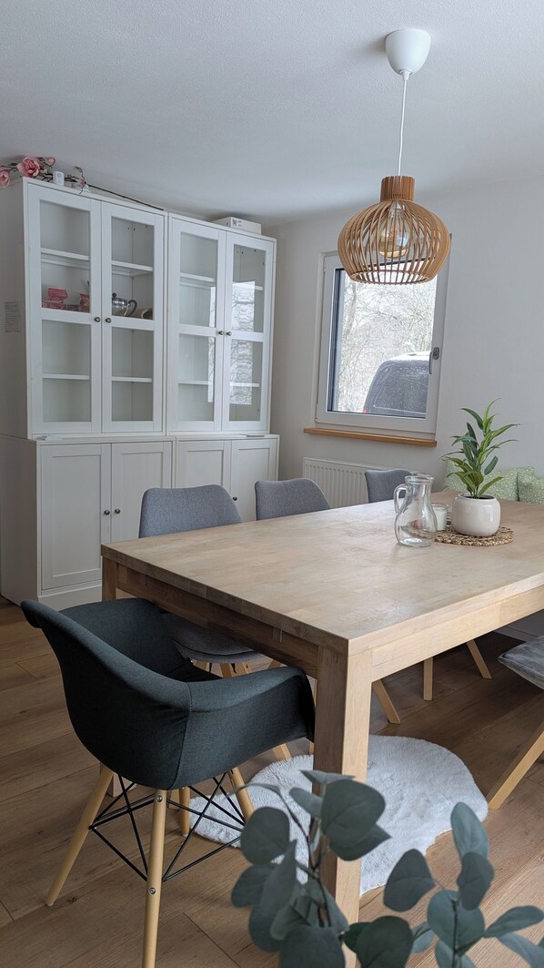 Un comedor con mesa de madera, sillas grises tapizadas, un armario blanco con puertas de cristal, una lámpara colgante y una ventana con vistas.