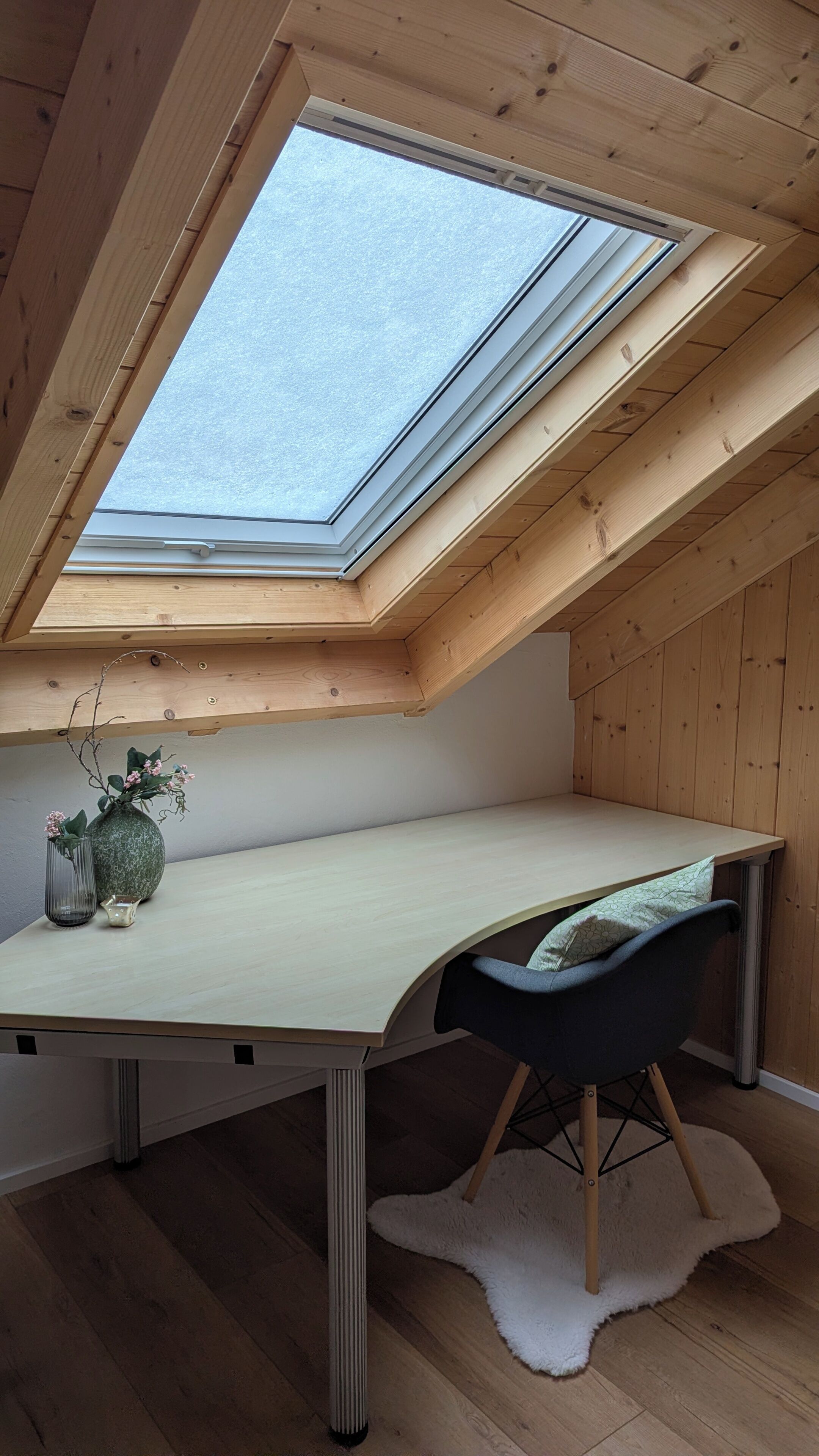 Une pièce avec un plafond en pente, un puits de lumière, un bureau, une chaise et un petit vase avec des fleurs.