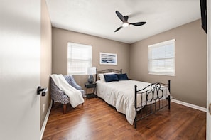 A bedroom with a bed, a chair, a nightstand, a ceiling fan, and two windows.