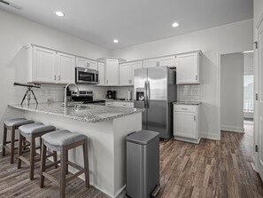 A modern kitchen with white cabinets, stainless steel appliances, a granite countertop, and wooden bar stools. - Boater's Cove, 3BR/2BA, Neighborhood Boat Ramp (Cedar Point)