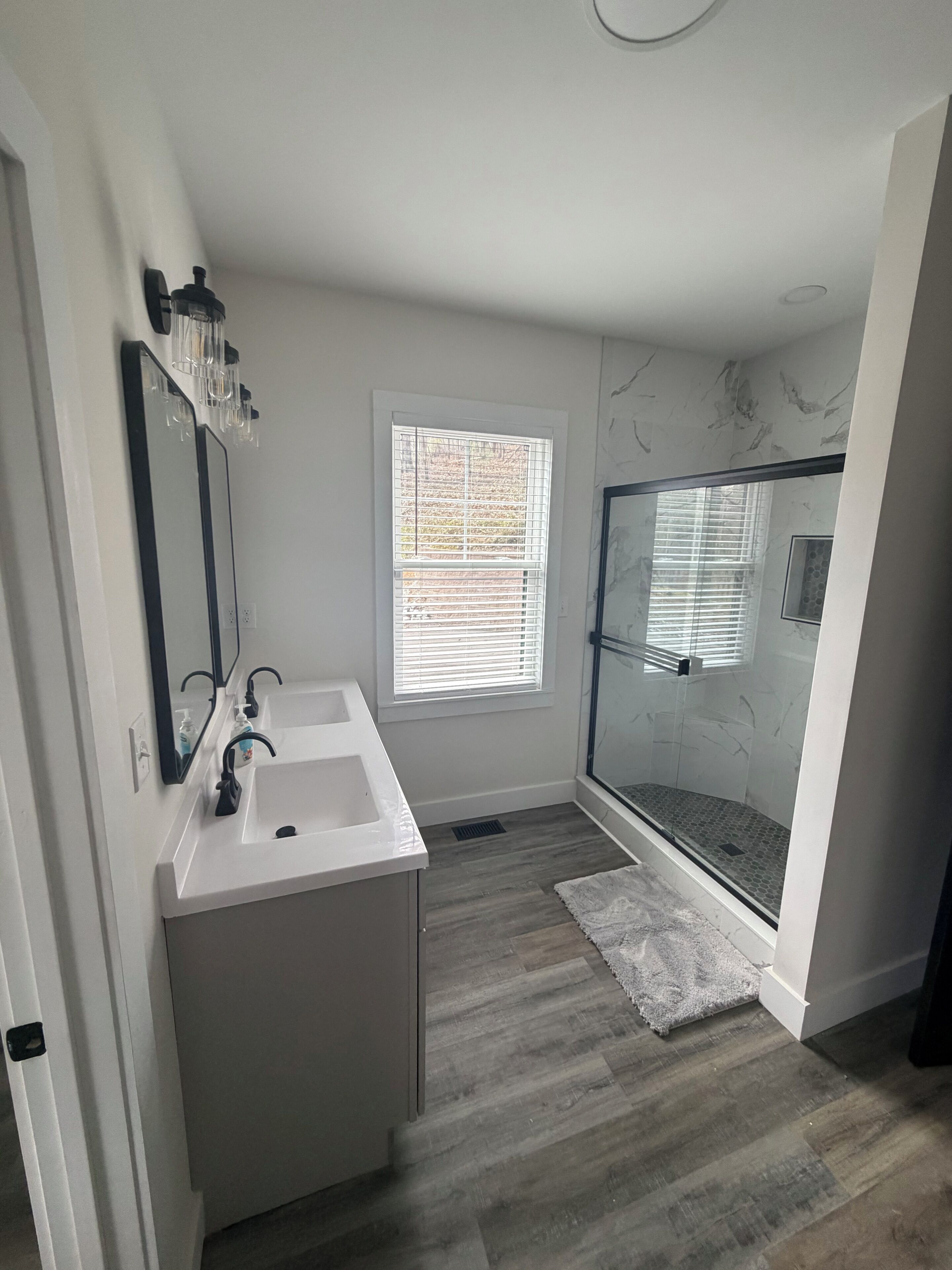 Une salle de bain avec un meuble-lavabo blanc, un grand miroir, une douche avec des portes en verre et une fenêtre avec des stores.