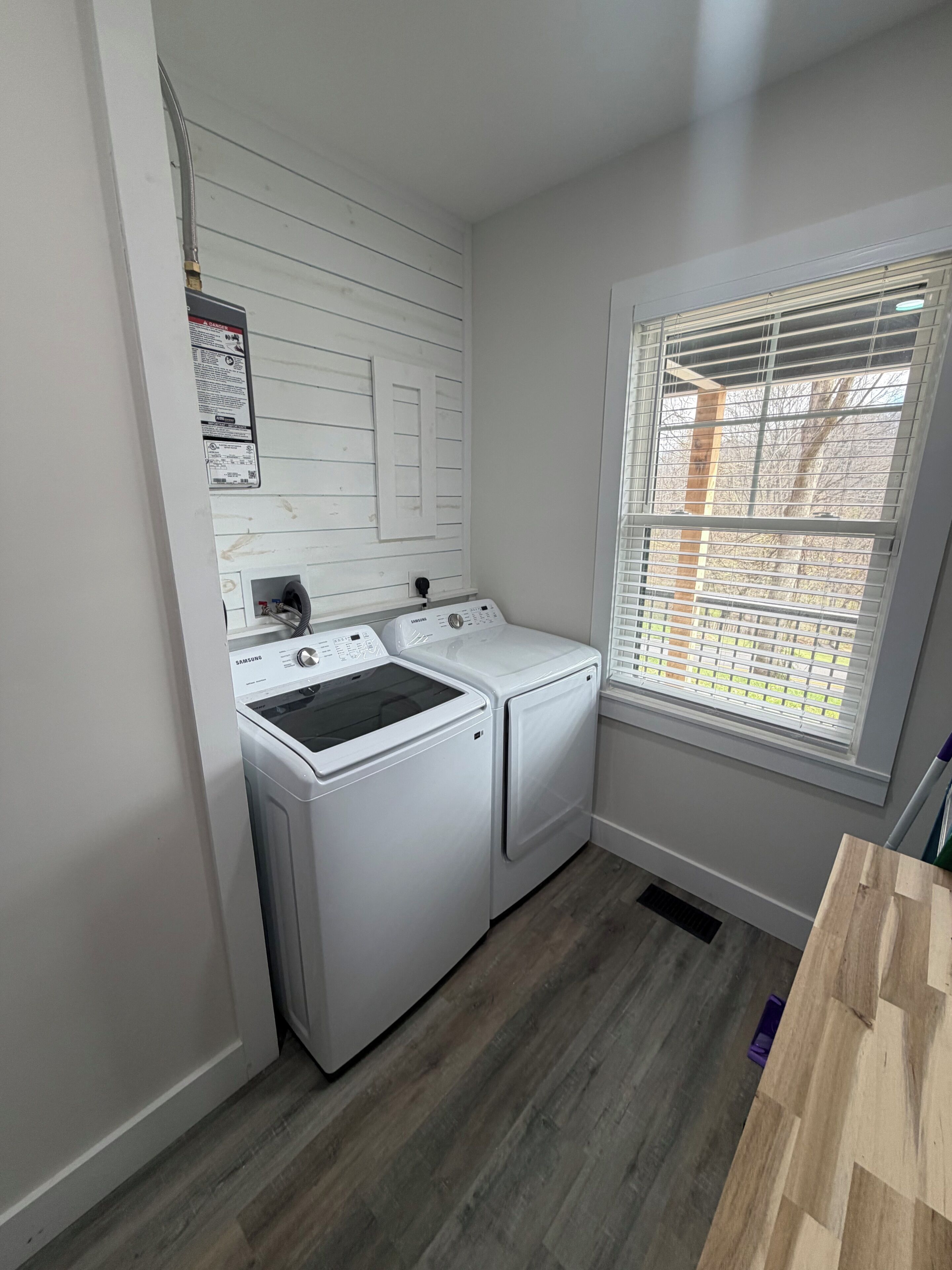 A laundry room with a washing machine and dryer, a window with blinds, and a wooden floor.