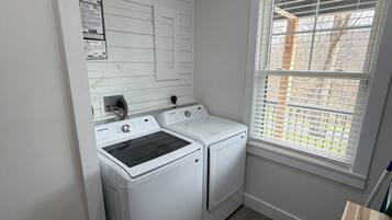 A laundry room with a washing machine and dryer, a window with blinds, and a wooden floor.