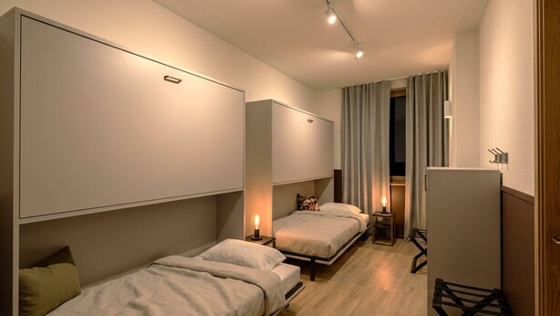 Une chambre d’hôtel compacte, équipée de deux lits, d’un petit bureau, d’un réfrigérateur et d’une fenêtre avec des rideaux.