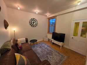 A living room with a brown sofa, a flat-screen TV, a decorative clock, and a window with blinds. - Stylish 3BDR Home Steps from Leytonstone Tube (Greater London)
