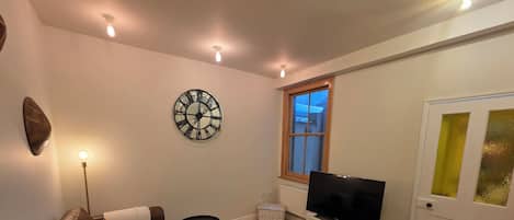 A living room with a brown leather sofa, a flat-screen TV on a white stand, a round wall clock, and a window with a view.