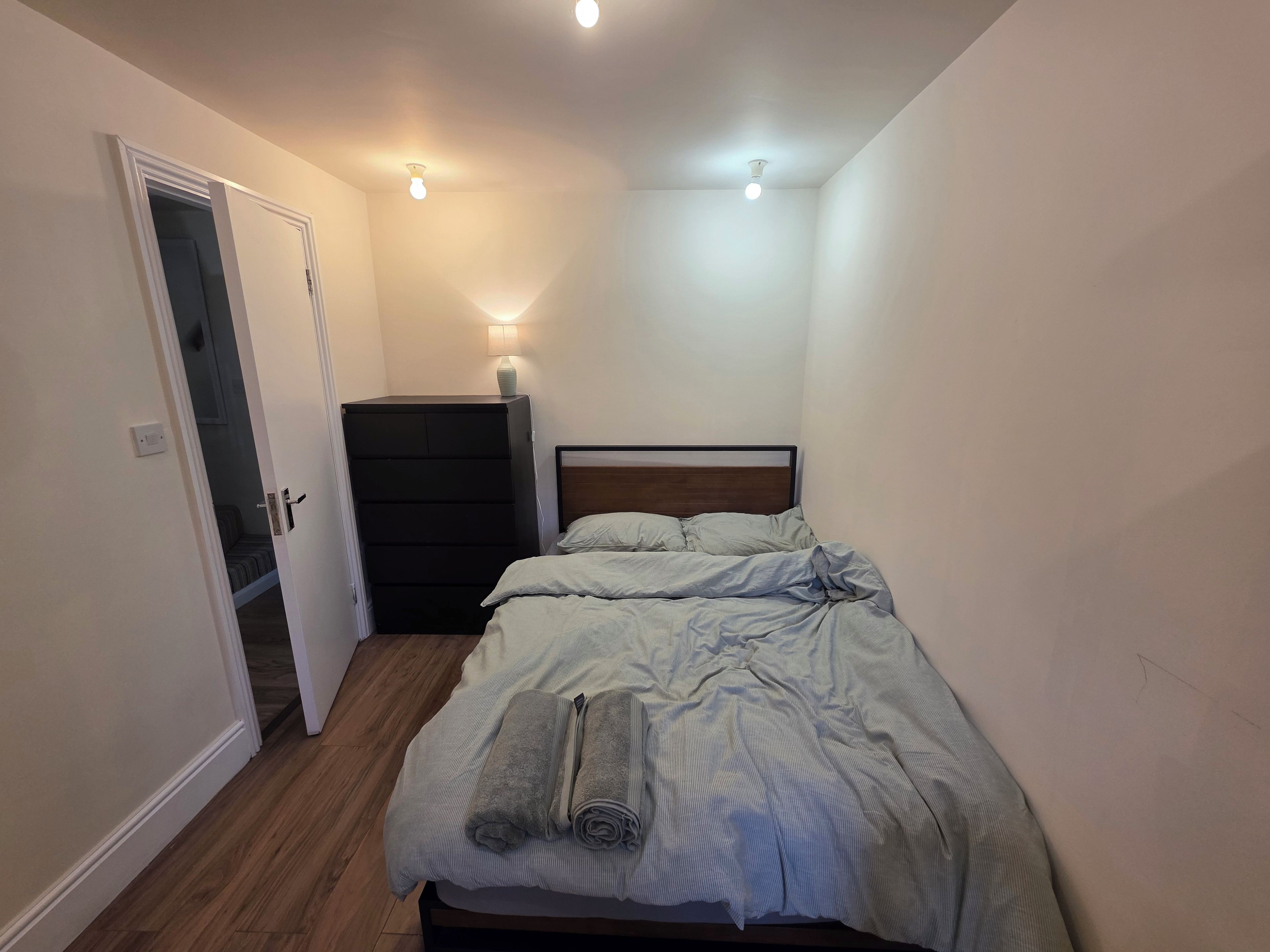 A bedroom with a bed, a dresser, a lamp, and a door leading to another room.