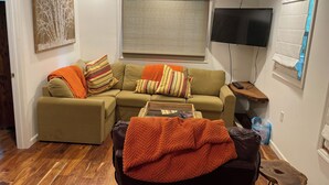 A living room with a sectional sofa, a coffee table, a ceiling fan, a TV, and a window with blinds. - Soothing 2-bedroom cabin with AC in charming Bassett (Bassett)