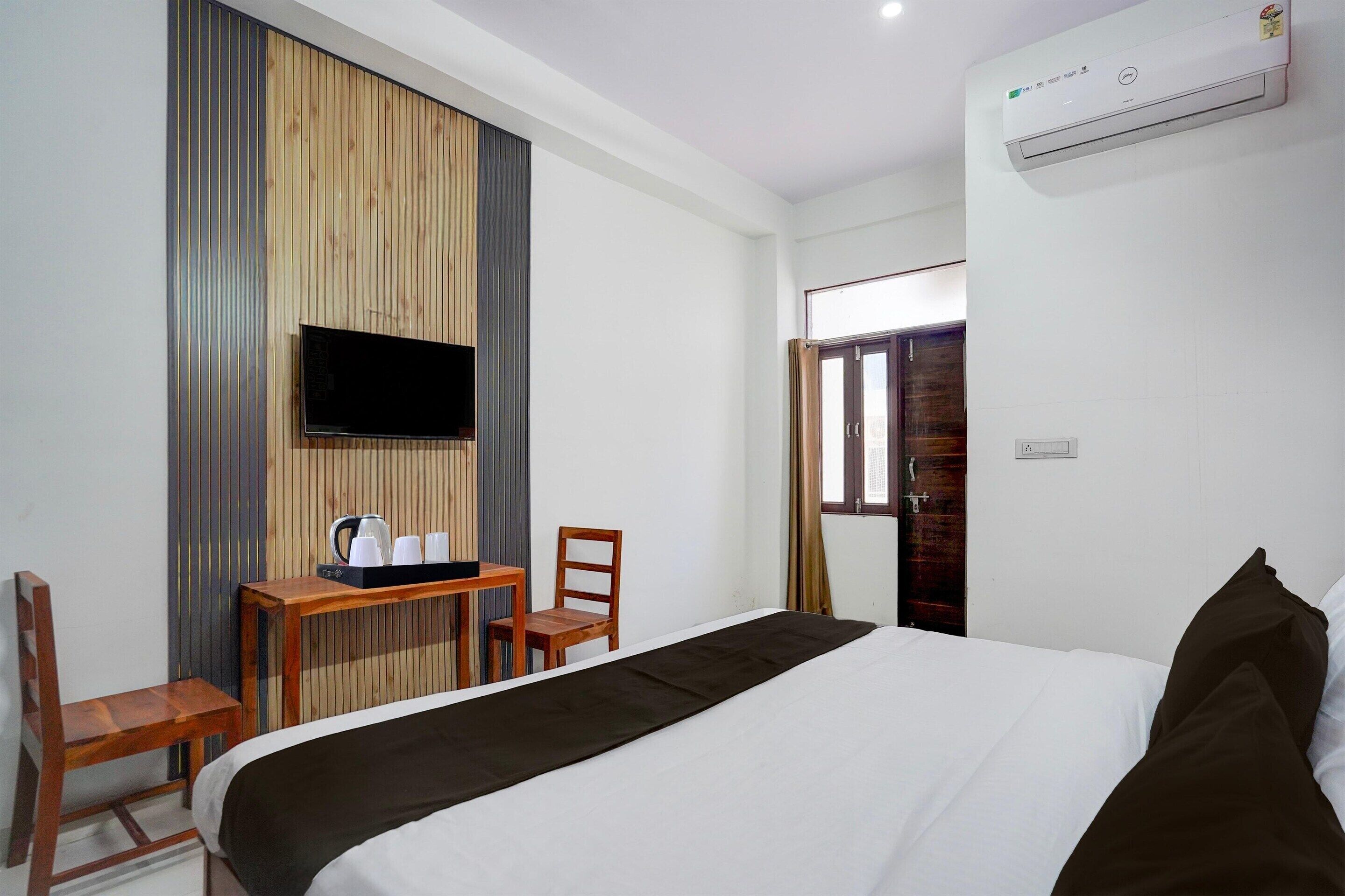 Una habitación de hotel con una cama, un escritorio con hervidor, una silla, un televisor montado en la pared y un aire acondicionado.