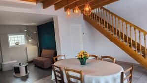 Un coin repas avec une table ronde, des chaises, un vase de fleurs, un banc, un radiateur, une fenêtre donnant sur un paysage et un escalier en bois.