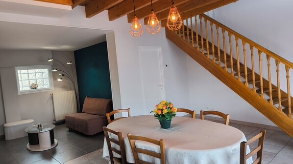 A dining area with a round table, chairs, a vase of flowers, a bench, a radiator, a window with a view, and a wooden staircase.